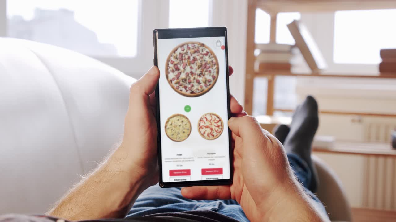 躺在沙发上的男人通过智能手机上的外卖应用在模糊的网上商店订购披萨。观点视频下载
