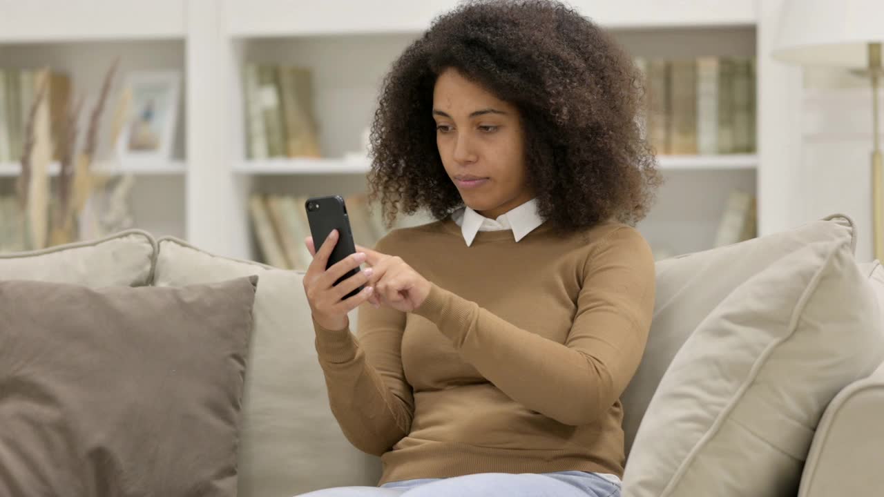 年轻的非洲女性对沙发上的智能手机的损失的反应视频素材