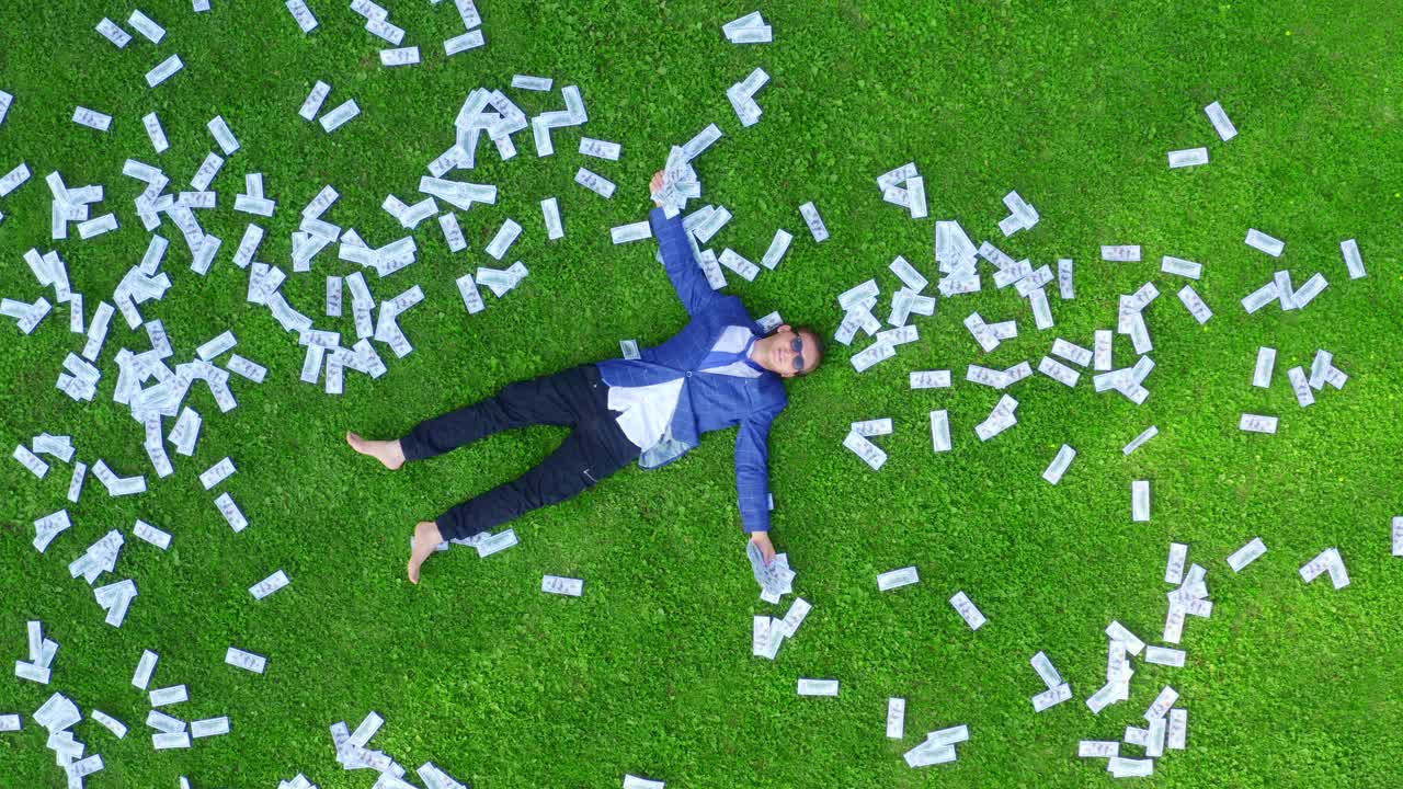 年轻的商人、学生或上班族躺在铺满钱的绿草地上。视频素材