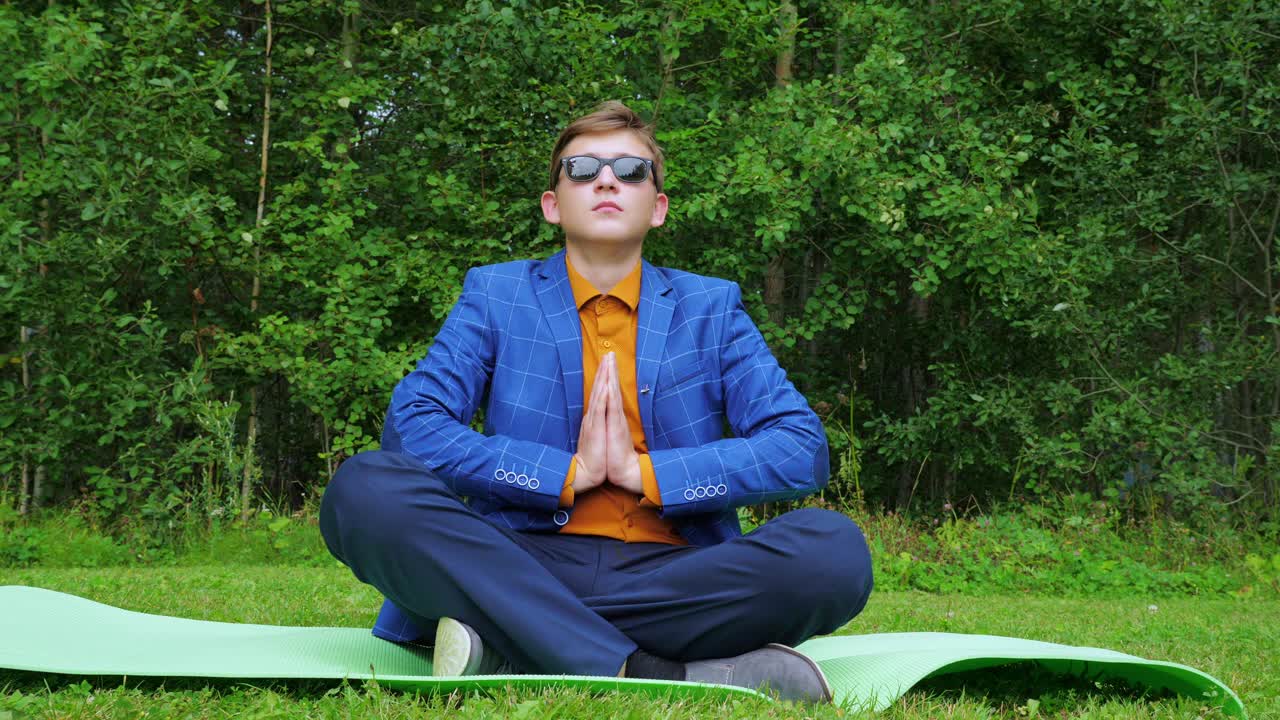 一个年轻人，一个十几岁的年轻人，穿着商务套装，坐在公园的训练垫上练习瑜伽。视频素材