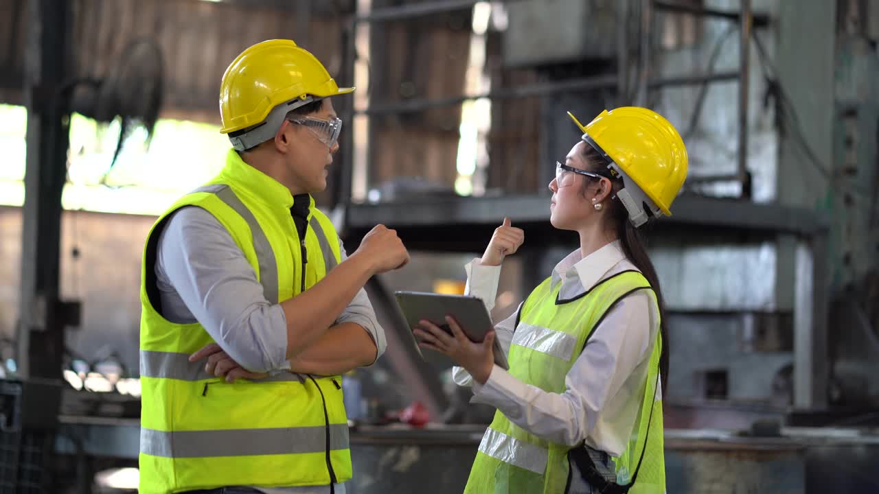 亚洲工业工程师经理男戴着眼镜和头盔与机工女讨论，同时用数字平板检查工业制造的大型工厂。检查视频下载