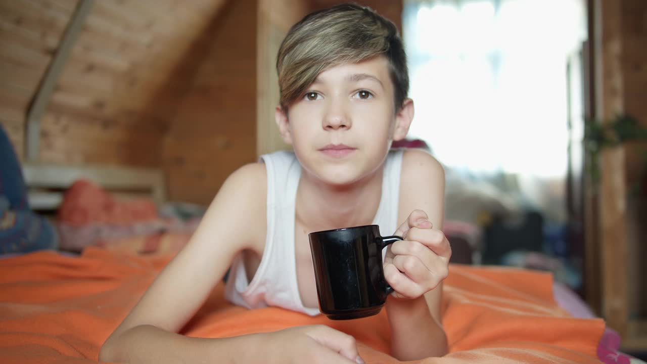 快乐的男孩躺在床上喝茶，兴致勃勃地看，乡村别墅，旅游视频素材