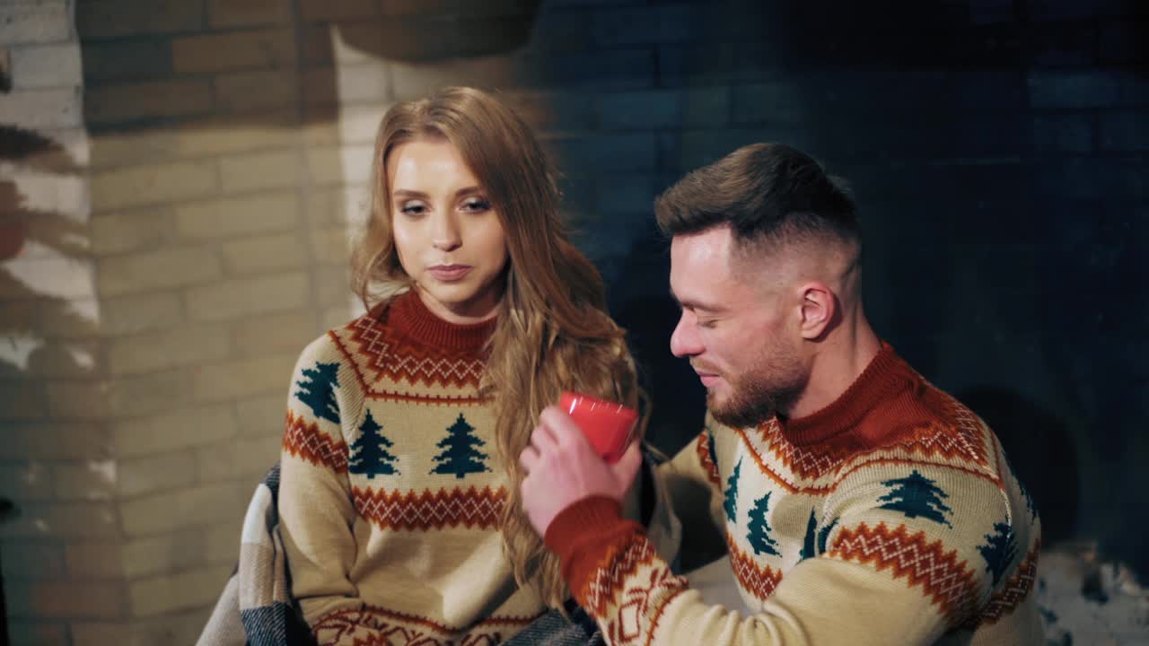 相爱的夫妇在喝茶。一个年轻人和一个漂亮的女孩穿着编织的圣诞毛衣坐在壁炉旁边。男人亲吻他的女朋友。浪漫的夜晚。视频素材