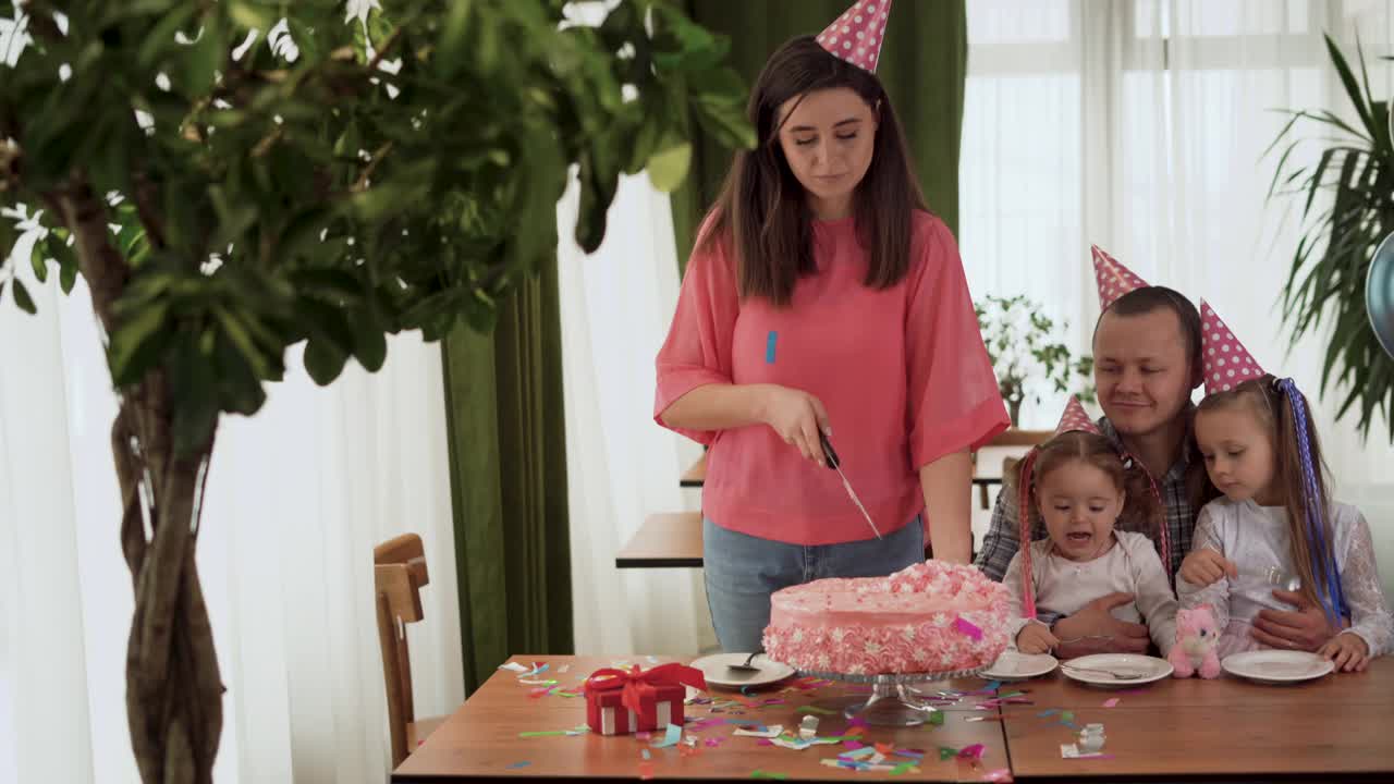 大家庭的爸爸，妈妈和两个女儿在家里庆祝女孩的生日。坐在家里的桌子旁，一个女人正在切生日蛋糕。视频素材