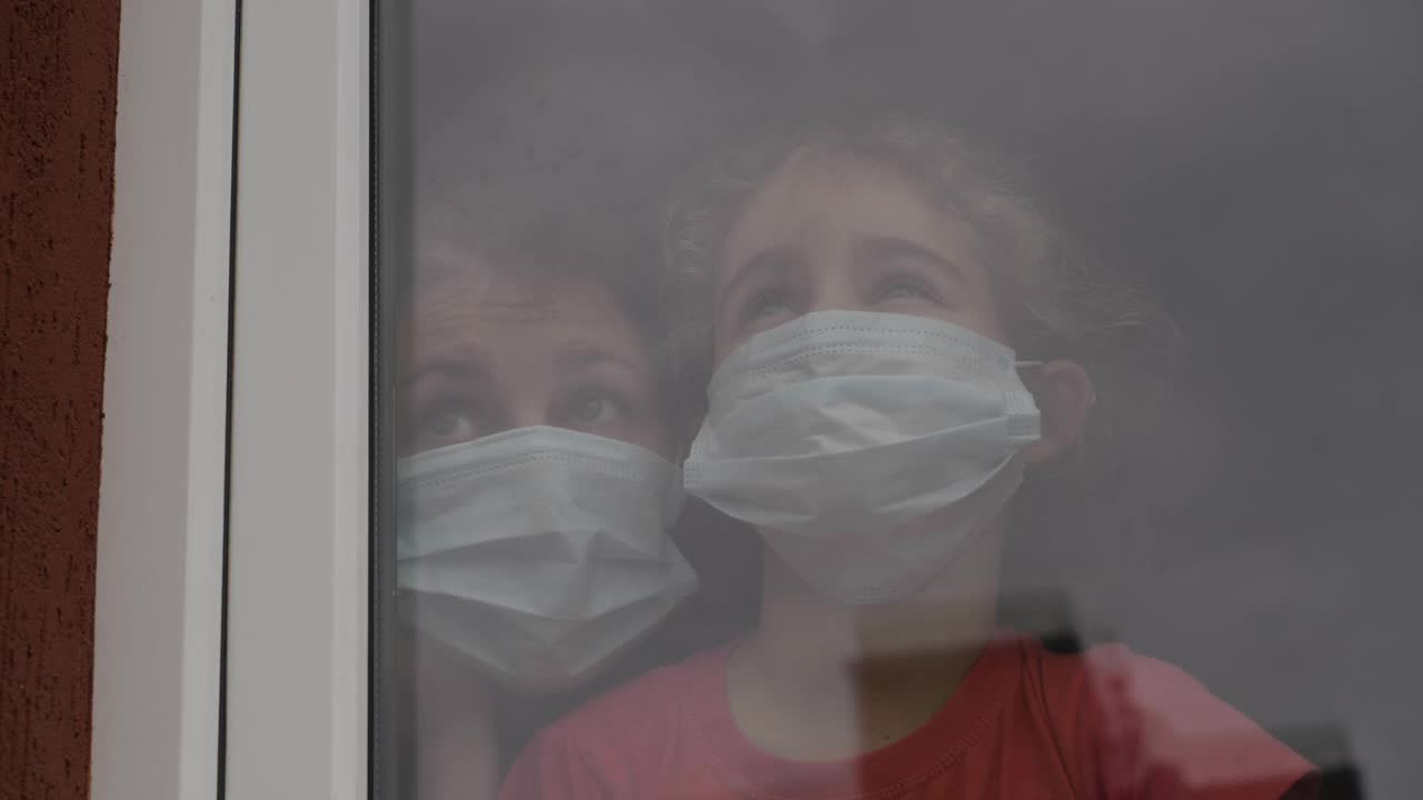 一名带着女儿的妇女，戴着防护口罩，悲伤地透过窗户看着，担心Covid-19被封锁。戴着防护面具的家人从窗户往外看。隔离冠状病毒社交距离视频素材