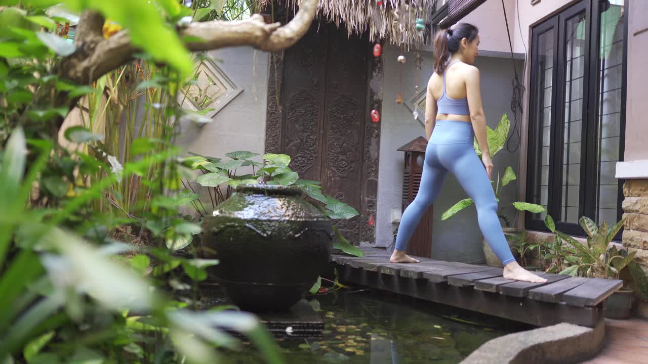 一位亚洲华人中年妇女在池塘上方的木桥上的前院练习瑜伽视频下载