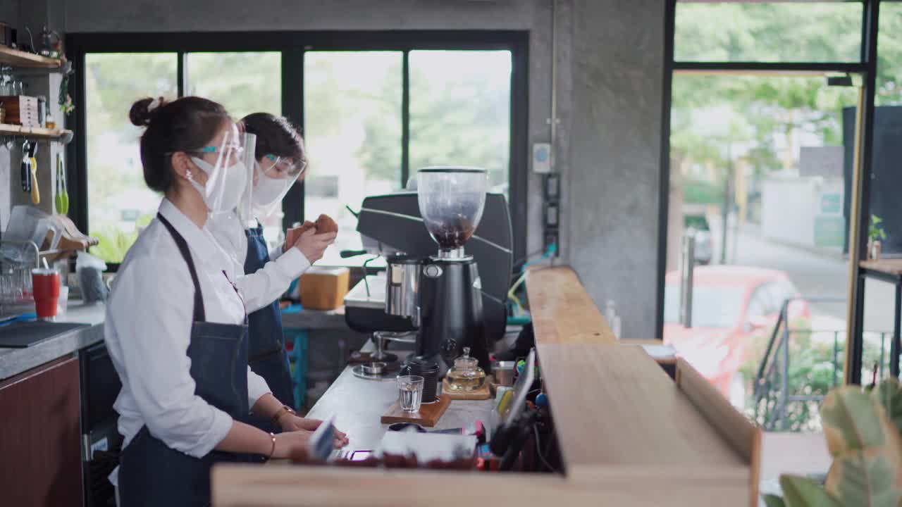 年轻的亚洲人café老板，女商人戴着口罩接受女性顾客的订单，使用手机扫描，并通过智能手机和非接触式数字平板电脑支付信用卡视频素材