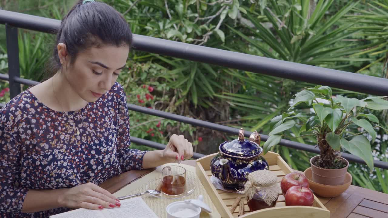 高角度的近景西班牙年轻女子搅拌热茶袋茶杯，而阅读在户外露台的一本书视频素材