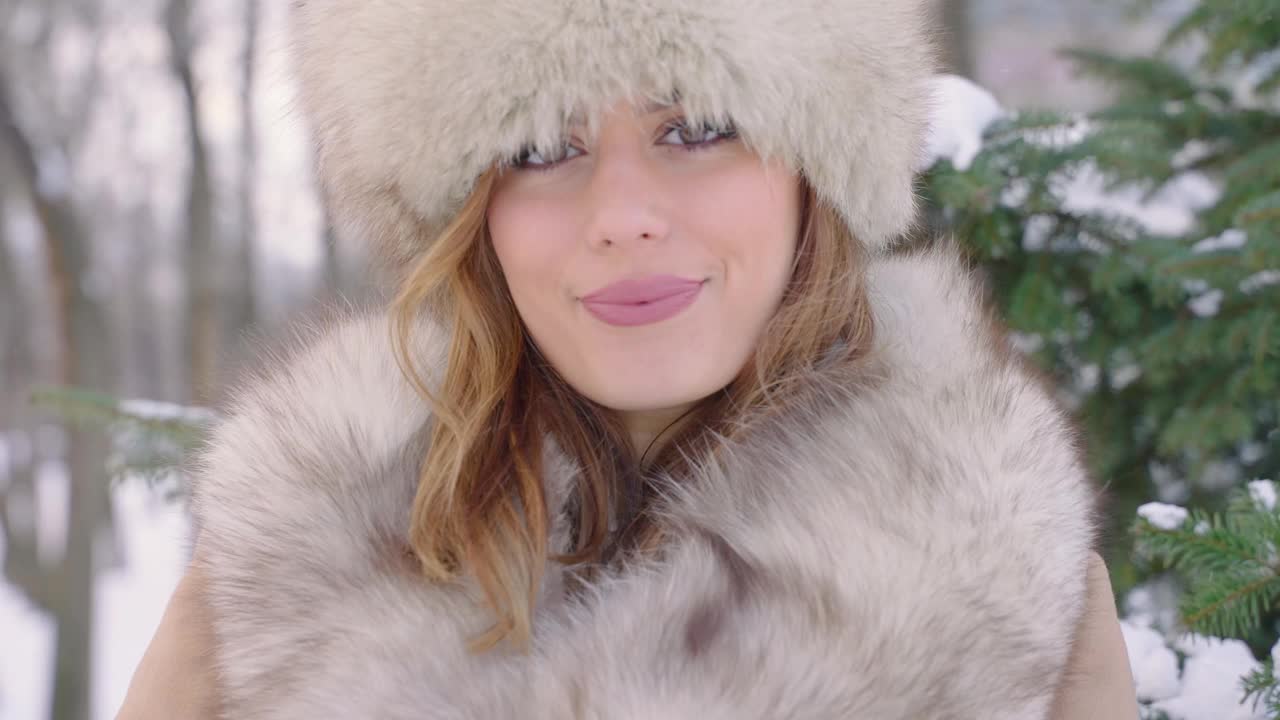 美丽的女人戴着皮帽看着相机在冬天的城市公园里白天在下雪的天气下飘落雪花，慢镜头视频素材