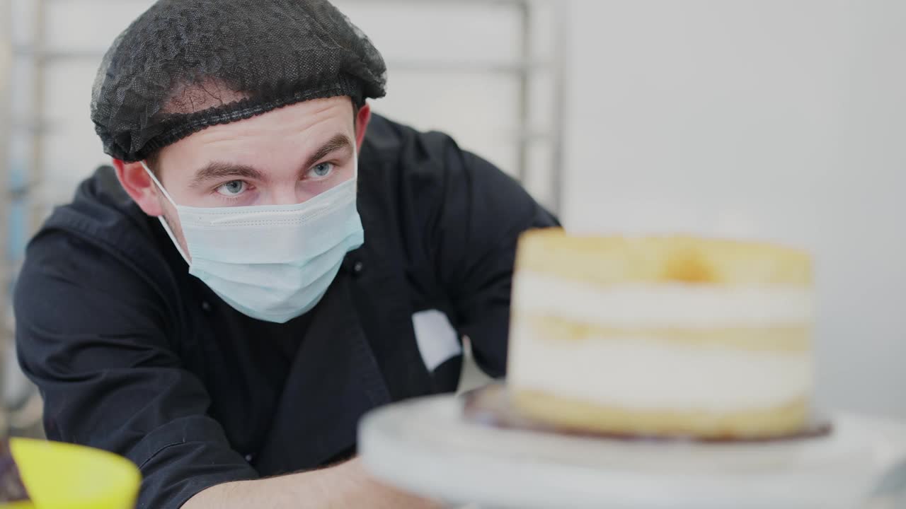 特写肖像集中厨师检查层蛋糕在旋转的桌子。一名严肃的白种人戴着冠状病毒口罩，看着糖果店厨房里美味的甜点。视频下载