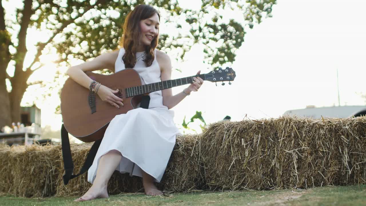 正面视图美丽的年轻亚洲女子在白色的衣服是弹奏吉他在一个草堆在早晨的阳光镜头光晕。视频下载