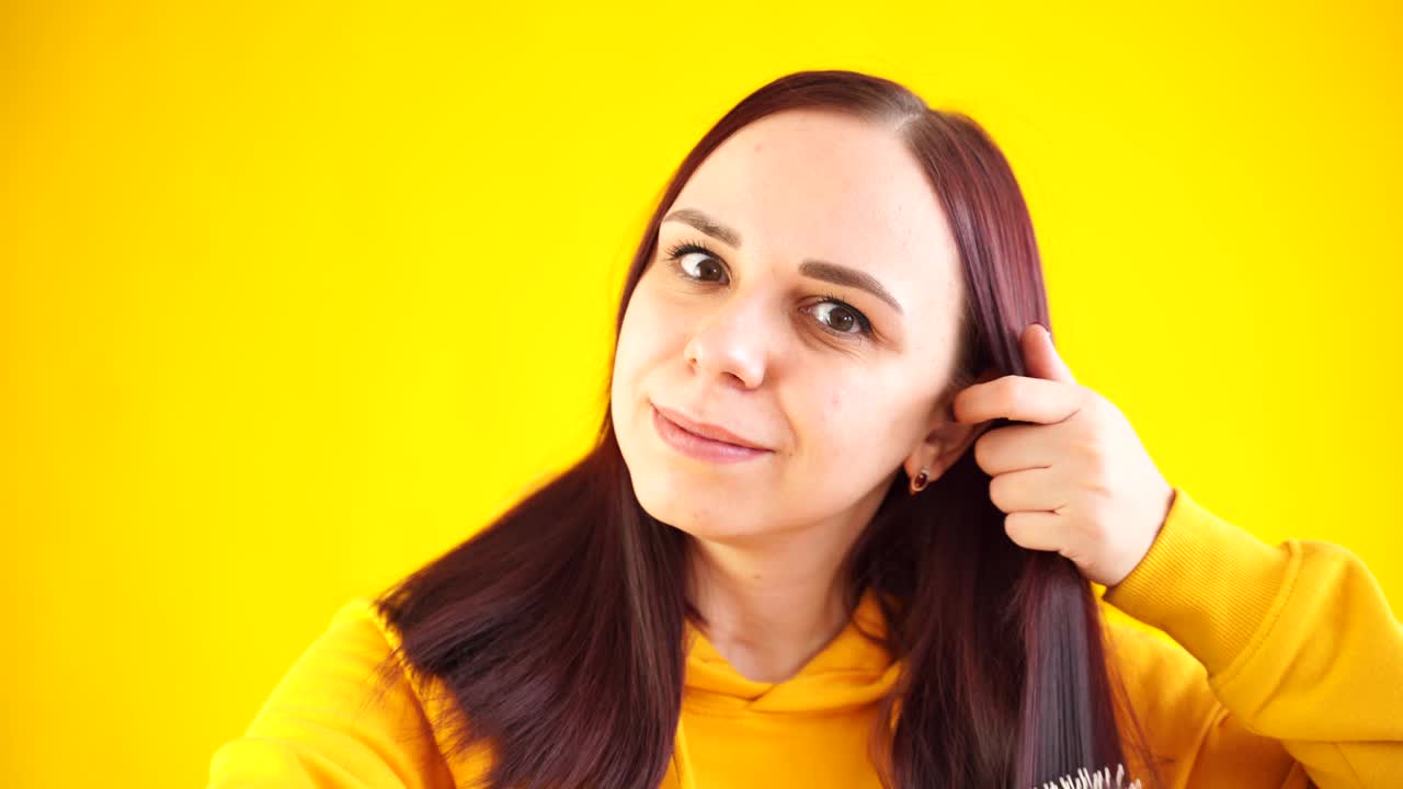 年轻女子的肖像梳理她的头发在黄色背景。穿着黄色连帽衫的褐发女子一边看镜头一边用嘴梳理视频下载
