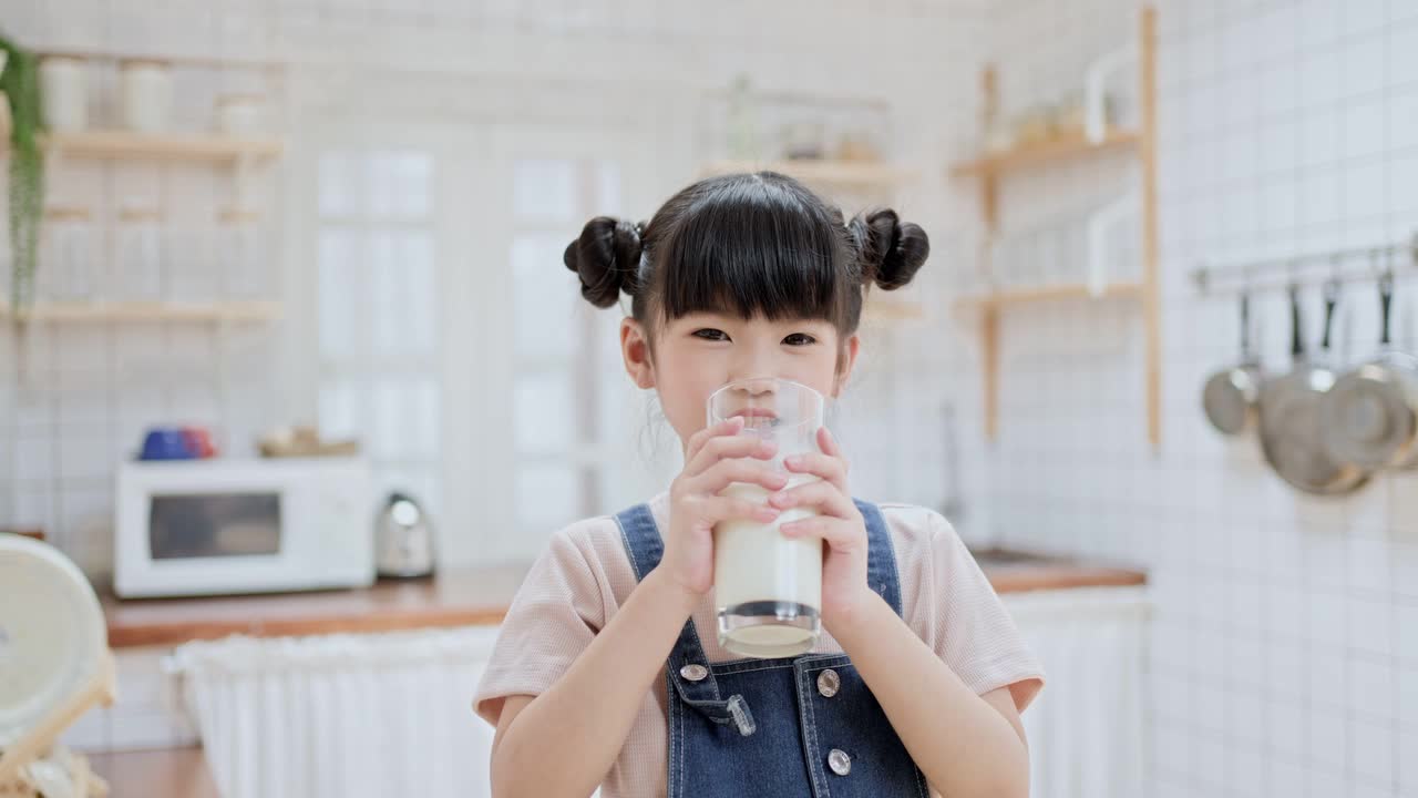 亚洲可爱的小孩拿着一杯牛奶在家里的厨房里喝。学龄前儿童或女儿被牛奶弄脏后，用手臂擦嘴，然后微笑着看镜头。视频下载