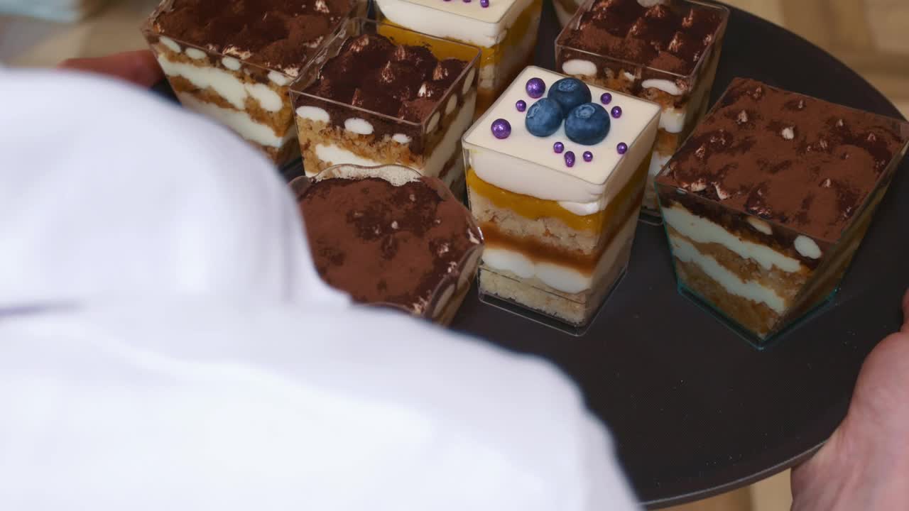 侍者端着一个棕色的托盘，上面放着各种以浆果、白巧克力和黑巧克力装饰的糕点，慢慢地走过天然木地板。在餐馆工作。特写镜头视频下载