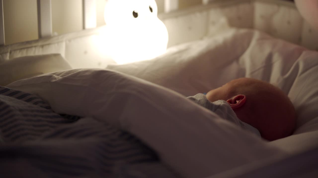 刚出生的男婴晚上睡在床上，3周大的婴儿在一盏夜灯下。视频下载