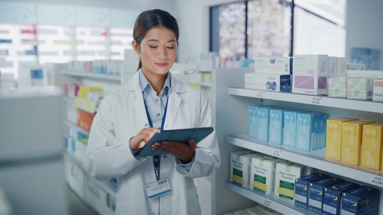 药店:美丽的亚洲药剂师使用数字平板电脑的肖像，看着相机和迷人的微笑，在她摆满药包的货架后面。中等的特写镜头视频素材