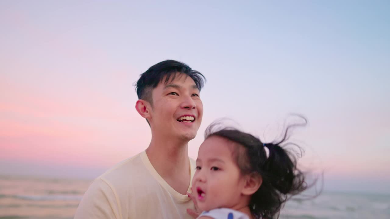 亚洲父亲抱着女儿在空中，而散步在海滩上的黄昏时间度假。视频下载