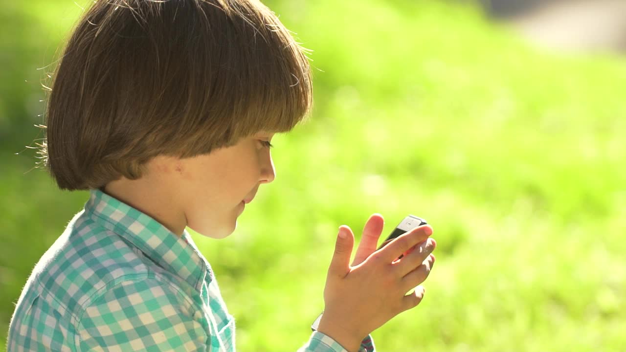 孩子玩手机在户外在绿色公园夏天。孩子使用wi - fi。可爱快乐的小男孩在智能手机上写短信，触摸屏，小玩意技术童年连接人的概念视频素材