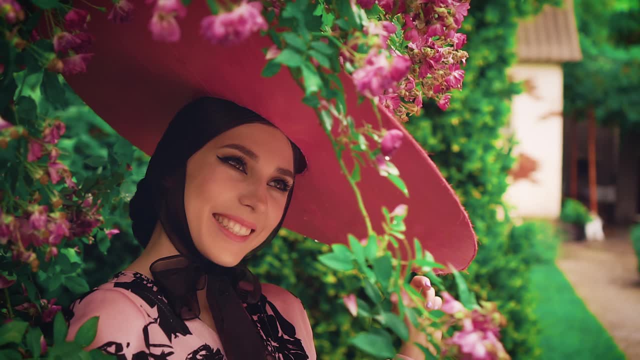 旧复古奥黛丽·赫本风格。粉红色的宽边帽子。自然化妆品概念，节日猫眼眼线化妆，可爱的脸性感的嘴唇。春天盛开的花园绿玫瑰丛中。时尚50 60年代视频下载