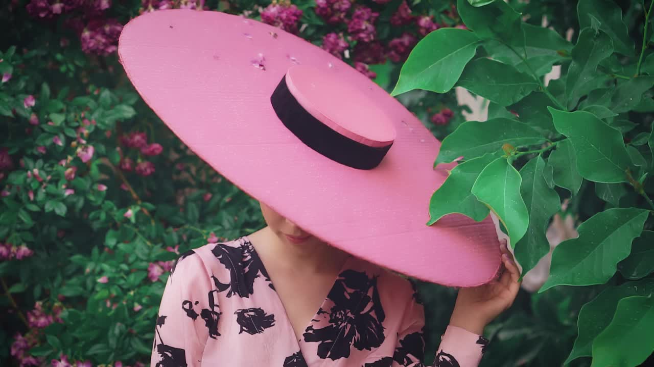 旧的复古风格。神秘的女士把脸藏在粉红色的宽帽下。自然化妆概念，节日猫眼眼线化妆，可爱的脸。春天盛开的花园绿玫瑰丛中。时尚50 60年代视频下载