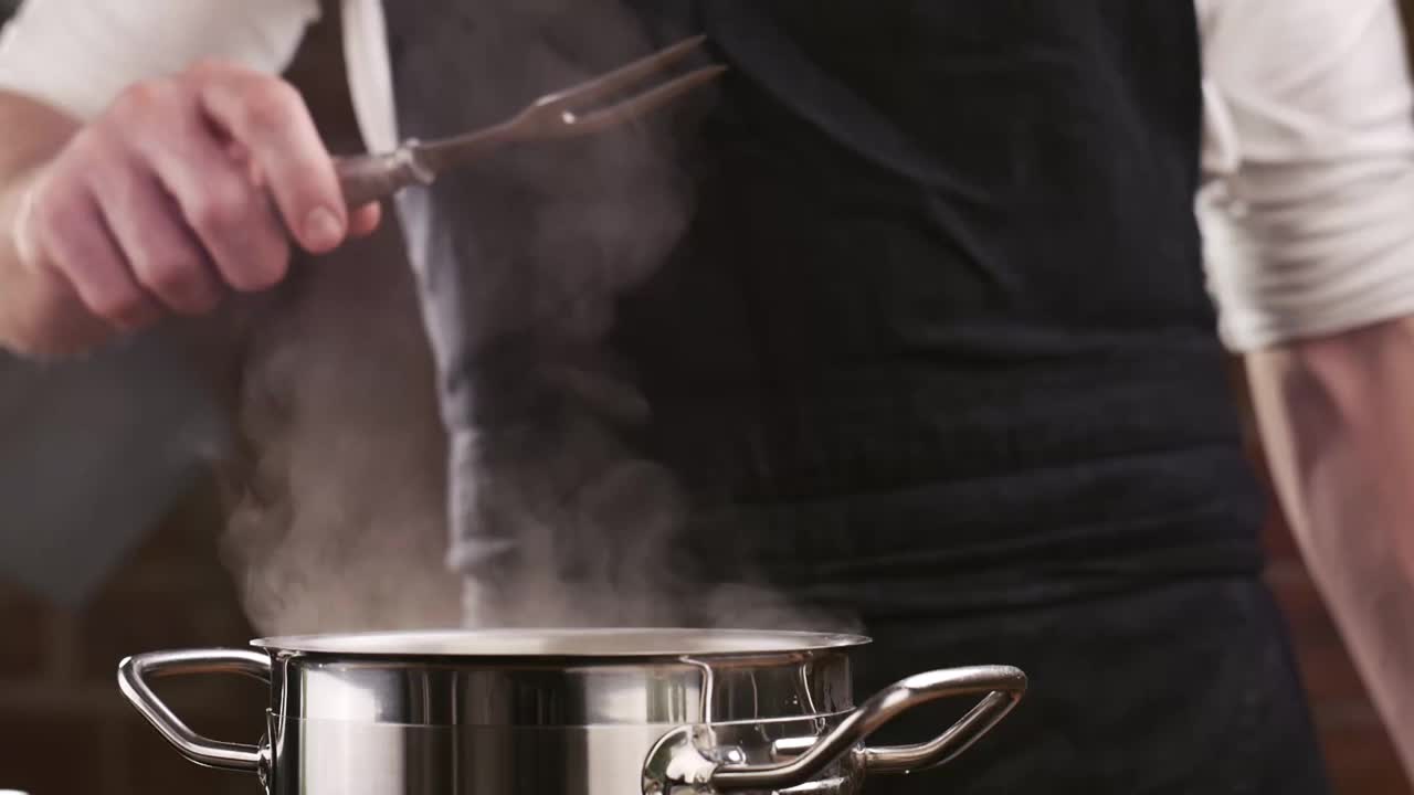穿着黑围裙的厨师从沸水中准备好意大利面。白烟以慢镜头从热锅上升起。在家做饭。美味的意大利面。视频素材