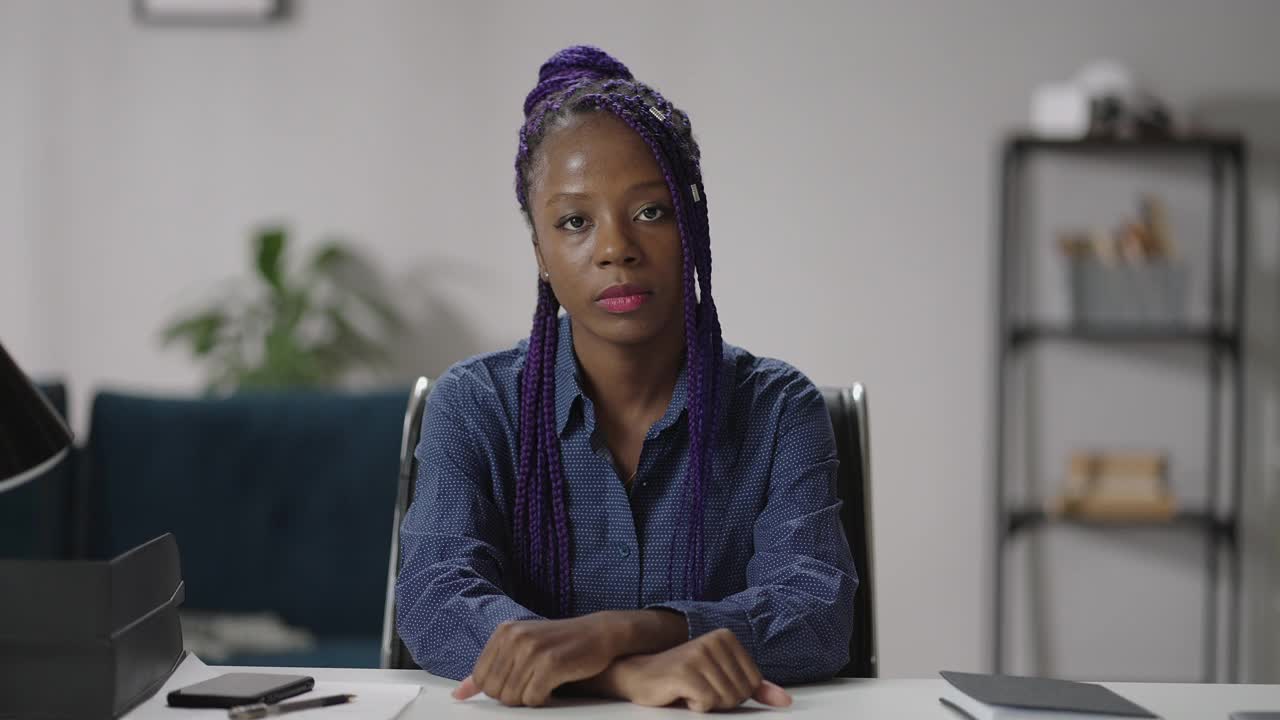 肖像严肃的非裔美国妇女，坐在桌子上，看着相机，微笑友好，在线聊天的概念视频素材