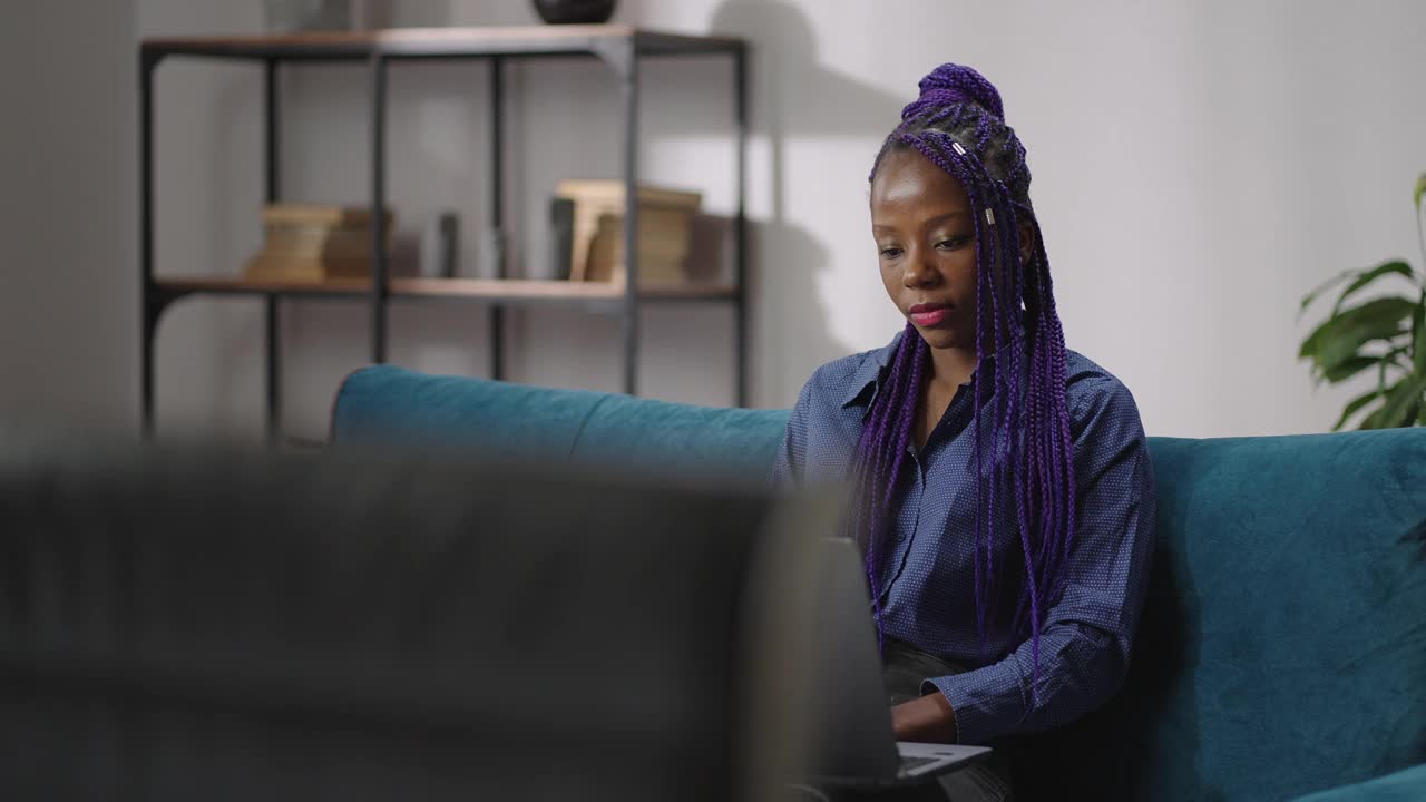 一位拿着笔记本电脑的美国黑人女性周末在家休息，坐在客厅的沙发上，通过视频电话进行交流视频素材