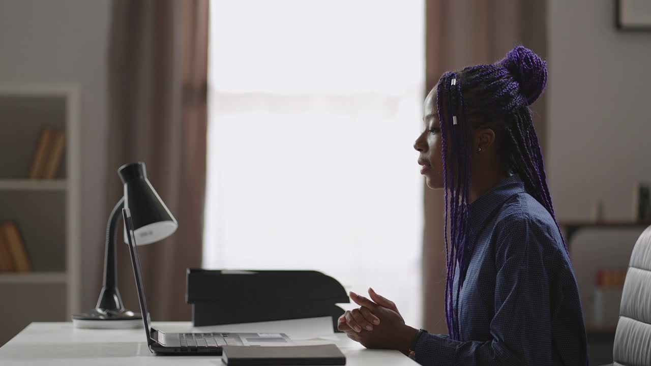 一名黑人妇女正在网上咨询，她坐在桌子旁，拿着笔记本电脑，看着网络摄像头，回答问题视频素材