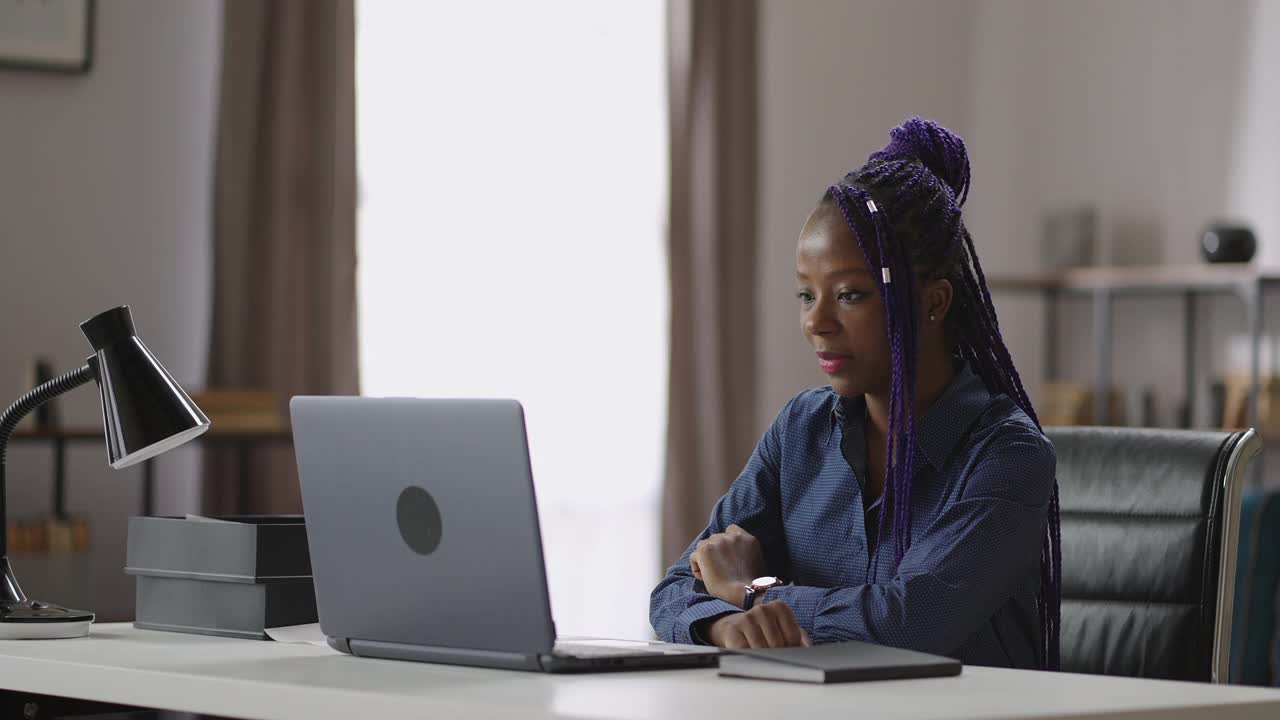 年轻的黑人妇女的肖像与紫色的长发绺在桌子上与笔记本电脑，非洲裔美国女士正在观看视频展示，在线会议或会议视频素材