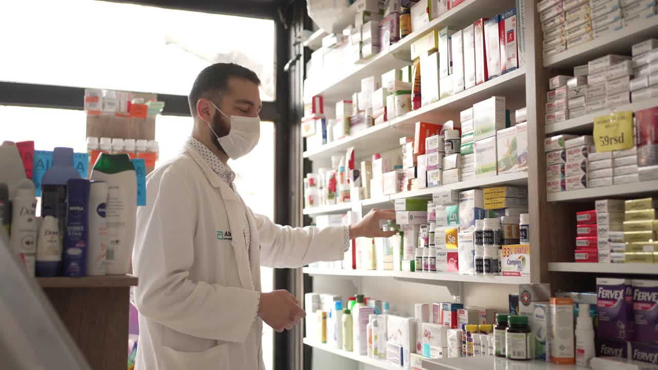 2019冠状病毒病大流行期间，男性药剂师戴着防护口罩在药店电脑上查看药品价格视频下载