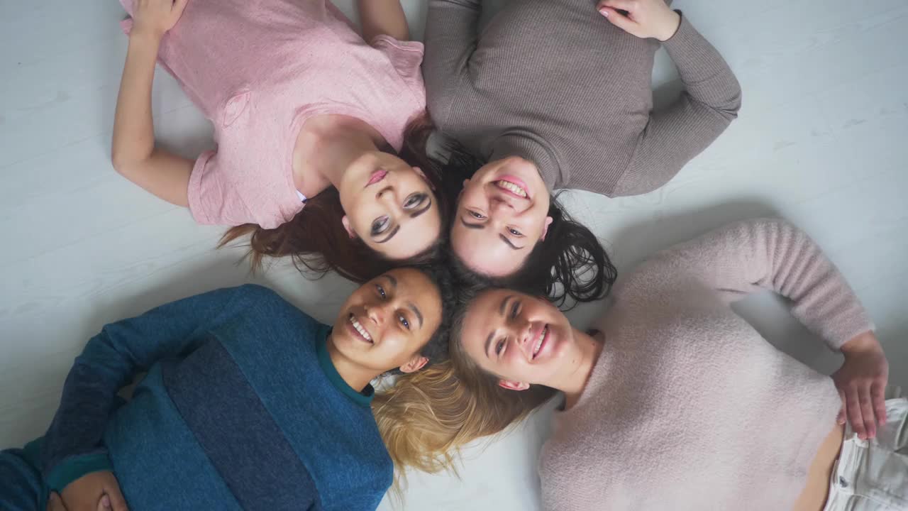 四个快乐的不同种族的漂亮的年轻女孩女性朋友躺在一起看相机，通宵派对和多民族的友谊概念，俯视图肖像视频下载