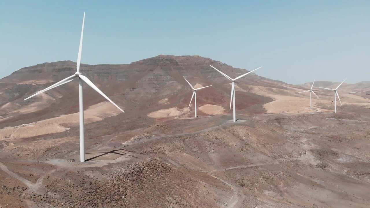 在阳光明媚的一天，富埃特文图拉岛沙漠景观中的风力涡轮机组的无人机镜头。能看到富埃特文图拉岛的街景。西班牙。视频下载