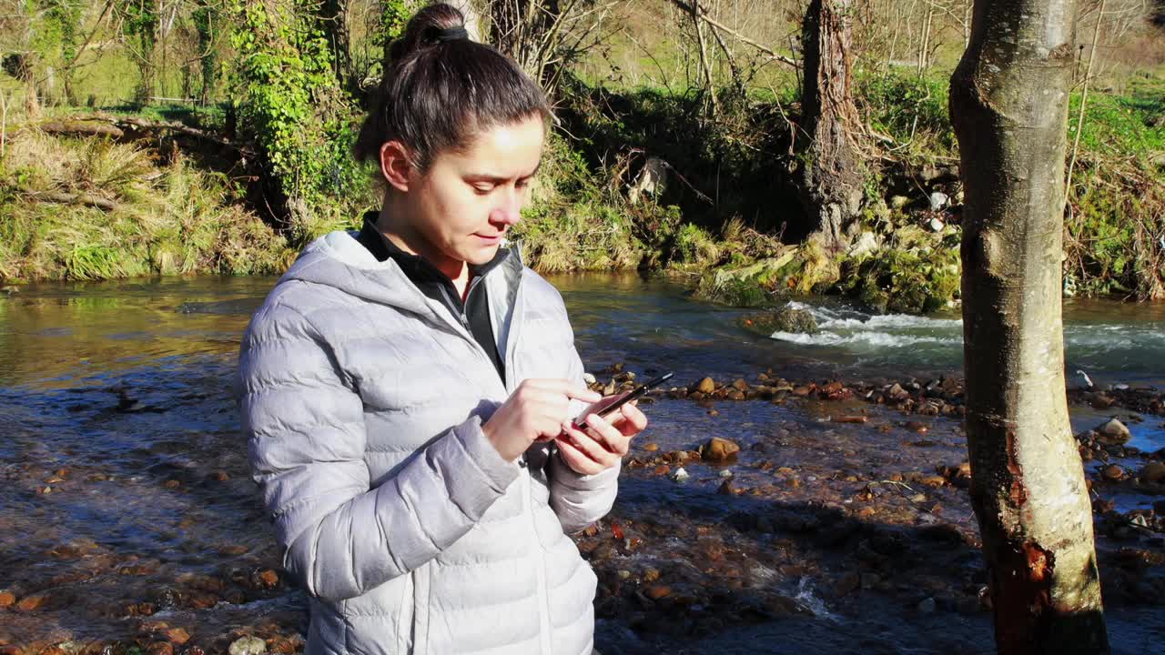 迷人的年轻女子在山里的河边用智能手机打字。视频下载