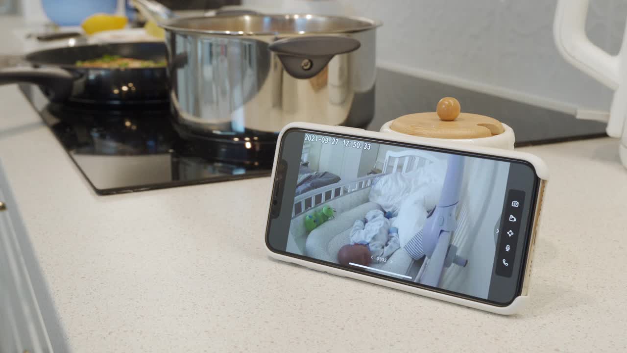 妈妈在厨房做饭，用ip摄像头作为手机上的婴儿视频监控，女人在智能手机屏幕上实时看着她熟睡的婴儿视频下载