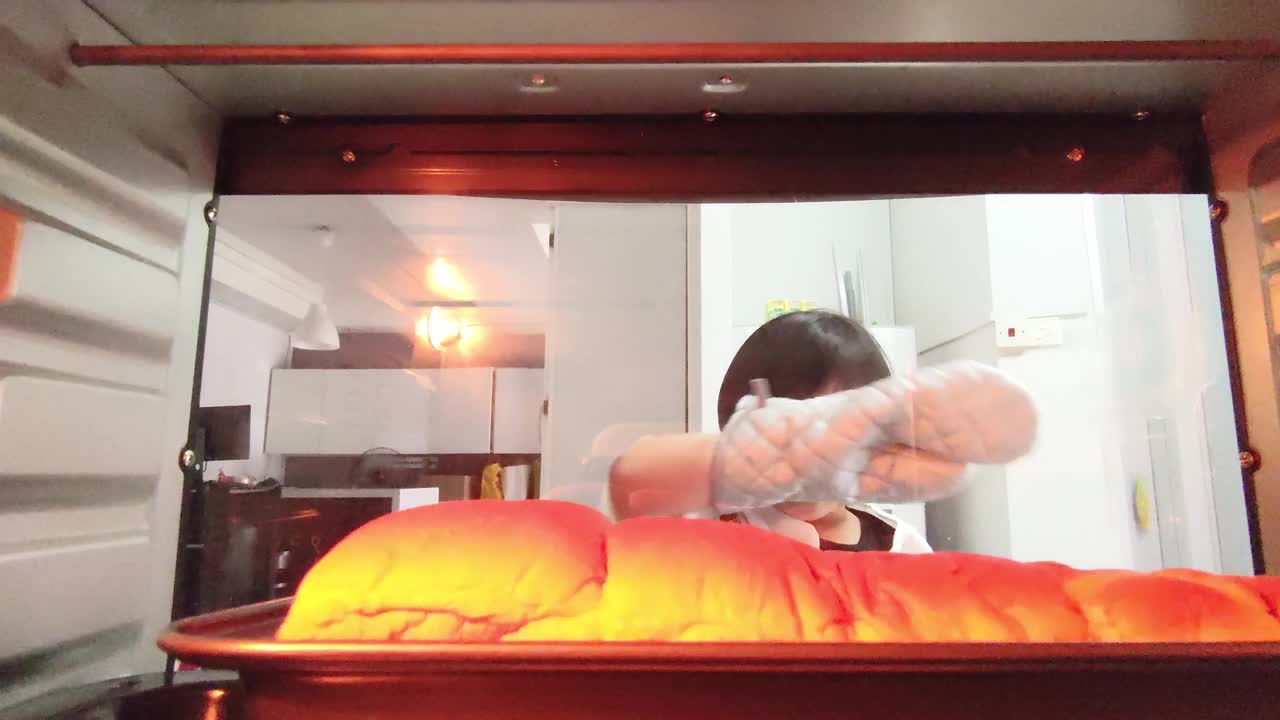 从烤箱里看，一个亚洲华人女孩在厨房里用防护手套从烤箱里愉快地取出烤好的面包视频下载
