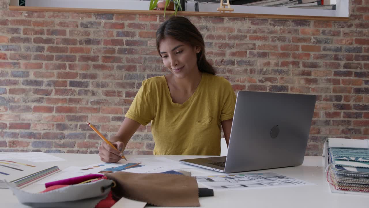 年轻的时装设计师在她的工作室里用她的笔记本电脑和不同的布料样板画草图，看起来很专注视频素材