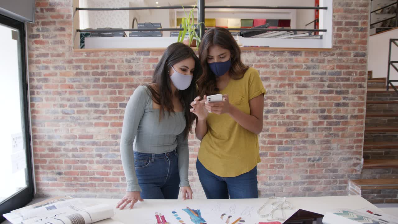美丽的时装设计师们在工作间隙用智能手机看社交媒体谈笑风生，两人都戴着防护口罩。视频素材