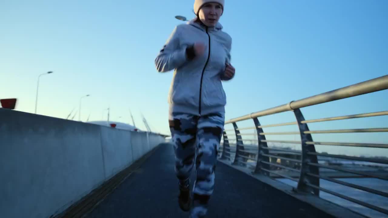 前面的摄像头记录了一个在桥上跑步的女孩穿着紧身迷彩服，白色运动衫，戴着粉色帽子。视频素材