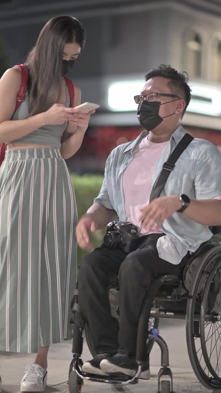 在吉隆坡，一名坐轮椅的亚洲华裔残疾男子与他的女性朋友在夜晚并肩交谈视频下载