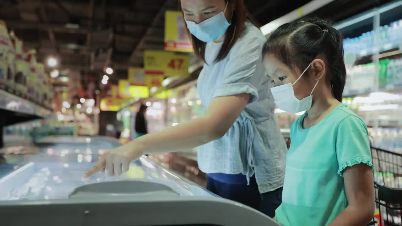 一名亚洲女童和她的母亲戴着防护口罩，以预防冠状病毒大流行，新的正常要求是一起选择超市冷藏室的冷冻食品。视频下载