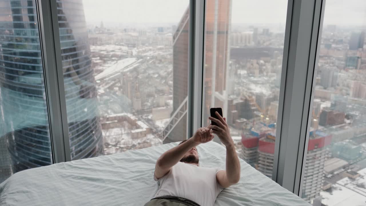 在一间现代化公寓里，一名男子躺在床上，手里拿着手机，旁边有一扇窗户，可以从顶层俯瞰这座城市，他通过视频连接进行交流视频下载