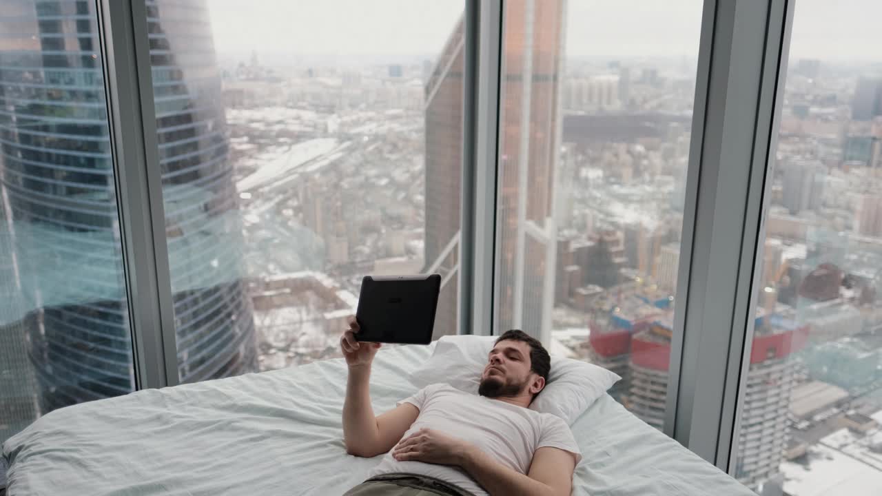 在一间现代化公寓里，一个年轻人躺在床上，手里拿着平板电脑，旁边有一扇窗户，从顶楼可以看到城市的景色视频下载