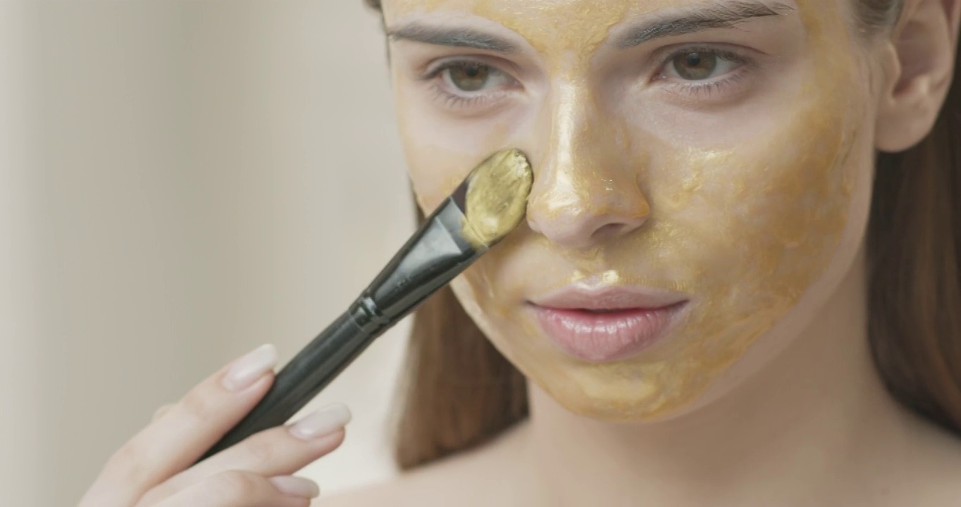 一个年轻的黑发女孩用刷子在皮肤上刷了一个金色的化妆面膜。视频素材