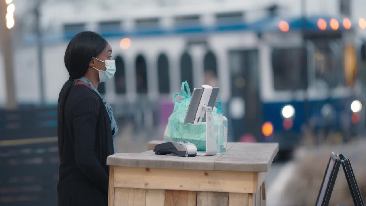 年轻的女服务员戴着面具，向一位接受路边订单的顾客挥手致意视频下载