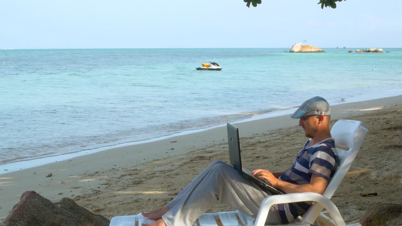 一名男子在沙滩上度假，在笔记本电脑上打字，享受着夏日的阳光视频素材