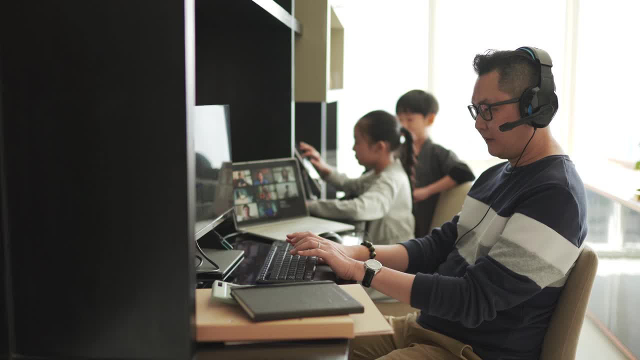 Lifehack亚洲华人父亲在家工作，用耳机与同事视频通话，而他的孩子在他旁边使用数字平板电脑视频素材