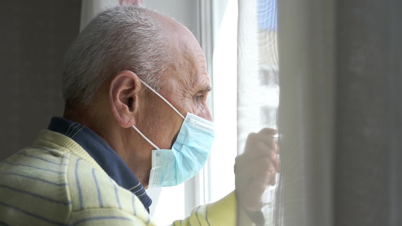 戴著蓝色消毒面罩的老人站在白窗旁视频素材