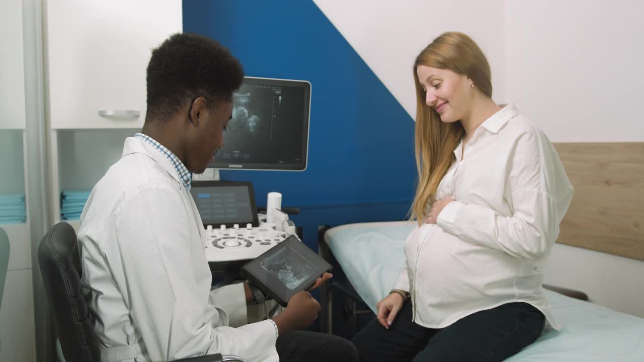 妇科会诊，超声概念。微笑的非洲男子医生产科医生显示数字平板与超声扫描的婴儿，以年轻快乐的孕妇在诊所视频素材