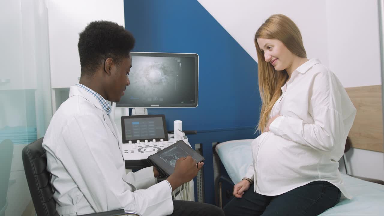 医生和病人在超声室。非洲一名男医生在对他的年轻孕妇进行超声波检查后，告诉她在平板上显示的超声波检查结果视频素材