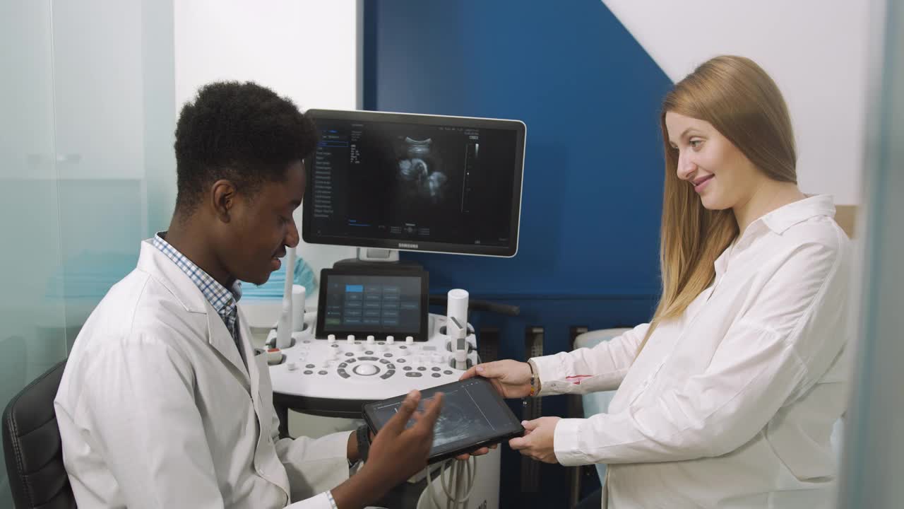 医生和病人在超声室。非洲一名男医生在对他的年轻孕妇进行超声波检查后，告诉她在平板上显示的超声波检查结果视频素材